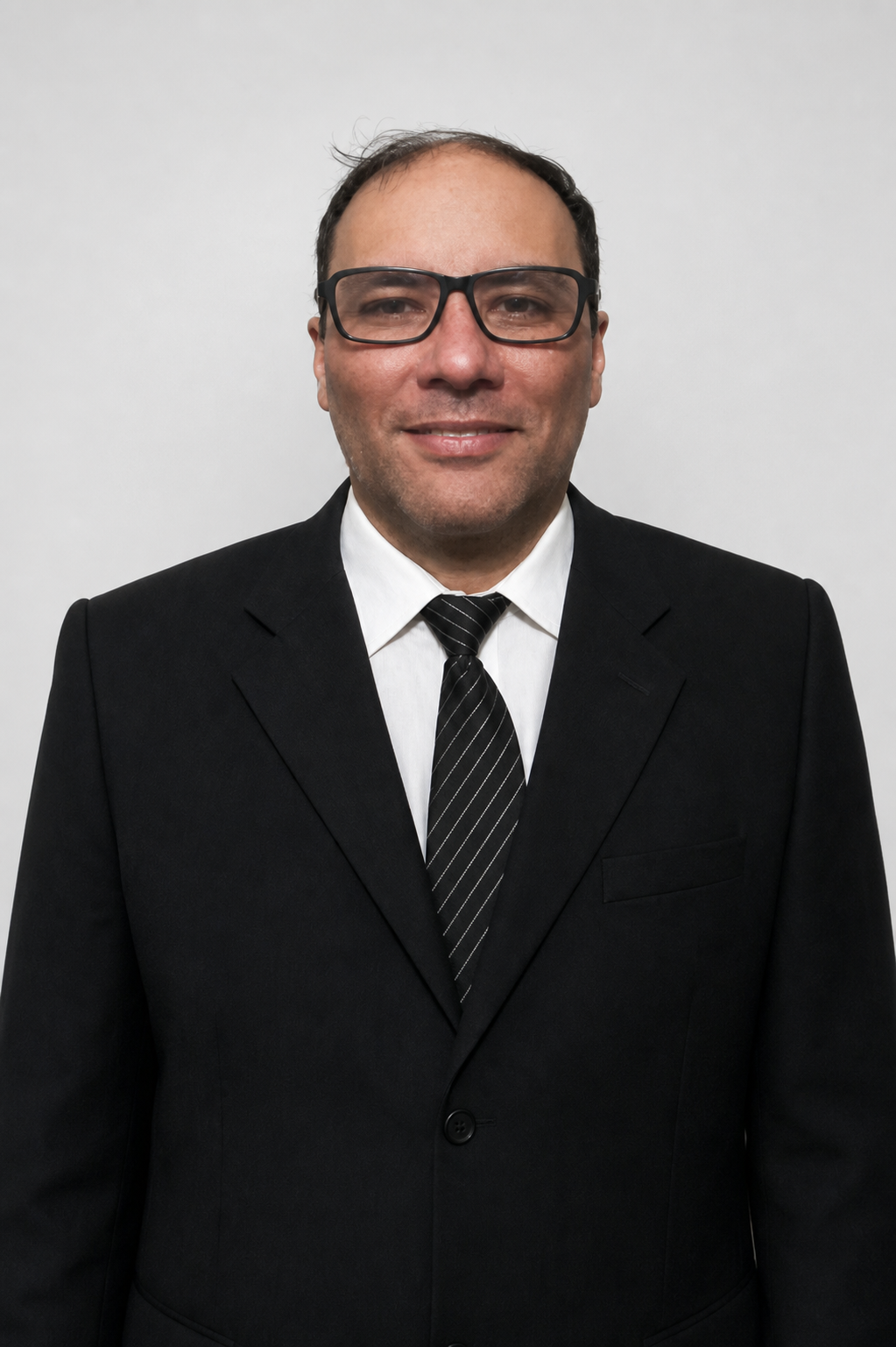 Fotografia restaurada de Mestre Mandíbula, Carlos Eduardo de Paiva Castro, usando óculos, terno preto, camisa branca e gravata listrada sobre fundo branco acinzentado